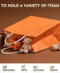 Orange kraft gift bag with handles holding plush toys and a gift box; Kraft Gift Bags With Handles Bulk Pack 24 Small Party Favor Bags