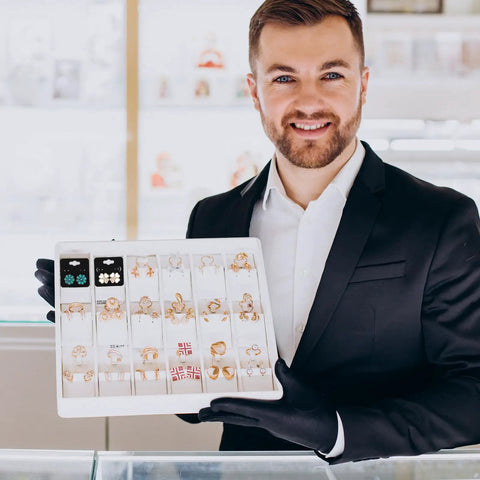 Jeweler holding display tray with various earrings, wearing black suit and gloves in a bright shop setting.
