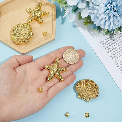 Slap These Killer Gold Cabinet Knobs Onto Your Dresser Or Vanity For An Instant Coastal Vibe Upgrade This Twelve Piece Hardware Set Features Detailed Starfish And Seashell Designs That Totally Revamp Your Space. A hand holding shiny gold starfish knob with a shell and fasteners on a tabletop.