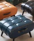 Brown, teal, and black leather ottomans with handle details on carpeted floor, showcasing tufted top and short legs for living room decor.