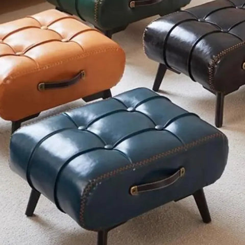Brown, teal, and black leather ottomans with handle details on carpeted floor, showcasing tufted top and short legs for living room decor.