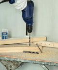 Blue handheld power drill with brad-point wood drill bits drilling wood; two spare bits on the workbench.