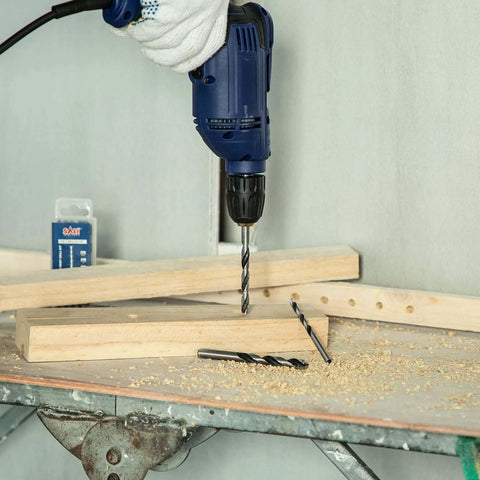 Blue handheld power drill with brad-point wood drill bits drilling wood; two spare bits on the workbench.