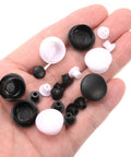 Hand holding black and white plastic snap-in screw hole plugs, various sizes, similar to highlighted 100 Pack Plastic Snap In Screw Hole Plugs 6mm Diameter Flush Shelf Button Caps For Furniture Cabinet Cupboard Dark Brown Durable Fits 5mm Under Head And 11.5mm Cap Diameter Keeps Surfaces Clean And Neat.