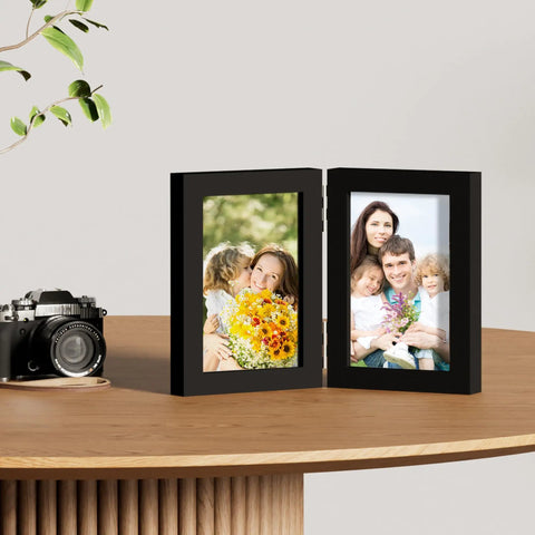 Double 4x6 Picture Frame Vertical From Engineered Wood With Plexiglass Bifold Photo Frame For Tabletop Display Holds Two 4x6 Photos In A Dope Black Finish That Fits Any Desk Or Shelf on wooden table with a camera.