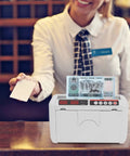 Portable Rechargeable Cash Counting Machine With UV And MG Counterfeit Detection on a counter with cashier in background