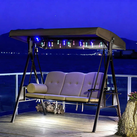Deluxe 3 Seat Backyard Outdoor Patio Porch Swing With Weather Resistant Steel Frame, Adjustable Tilt Canopy, Cushions And Pillow Included, Cozy Beige Upholstery, Supports Up To 750 Lbs For Three People illuminated at night on a deck.