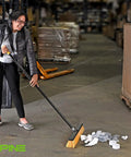 Real Deal Heavy Duty Push Broom With Long Handle Made To Crush Daily Grime On Smooth Floors Sweeps Sand Mud Leaves And Water Easy Without Breaking A Sweat In Bright Yellow 18 Inches Wide in use cleaning debris on warehouse floor.