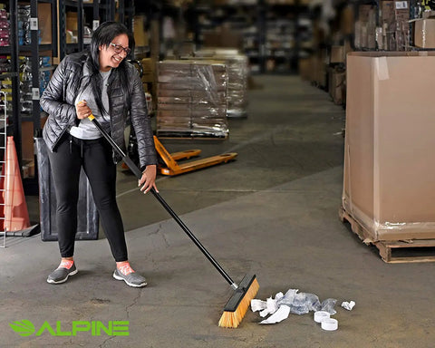 Real Deal Heavy Duty Push Broom With Long Handle Made To Crush Daily Grime On Smooth Floors Sweeps Sand Mud Leaves And Water Easy Without Breaking A Sweat In Bright Yellow 18 Inches Wide in use cleaning debris on warehouse floor.