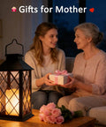 Woman and elderly lady exchanging gifts next to a 2-Pack 14-Inch Large Tempered Glass Solar Lanterns Outdoor Waterproof Hanging Lights With Realistic Flickering Flameless Candles For Wall Porch Patio Garden Cozy Decor Black on a living room table.
