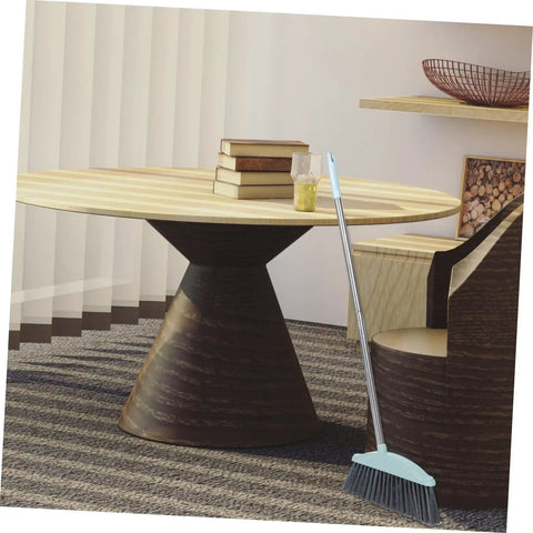 Grime Crushing Heavy Duty Broom With 34 Inch Long Handle For Sweeping Concrete Driveway Garage Patio And Indoor Floors No Sweat Household Cleaner With Convenient Storage Hook propped against a chair in a modern room.