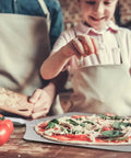 Thin Aluminum Pizza Peel With 13 And A Half Inch Wide By 16 Inch Long Surface And 8 Inch Wooden Handle For Indoor Oven And Outdoor Grillin Nonstick Transfer Paddle For Easy Pizza Bread And Pie in use with a child making pizza, tomatoes and spinach on table.