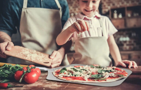 Thin Aluminum Pizza Peel With 13 And A Half Inch Wide By 16 Inch Long Surface And 8 Inch Wooden Handle For Indoor Oven And Outdoor Grillin Nonstick Transfer Paddle For Easy Pizza Bread And Pie in use with a child making pizza, tomatoes and spinach on table.