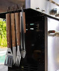 Stainless Steel BBQ Grill Tools Set with ergonomic wood handles hung on a grill, includes spatula, tongs, knife, and fork for outdoor grilling.