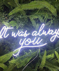 White neon sign with 'It was always you' phrase set against a green fern background.