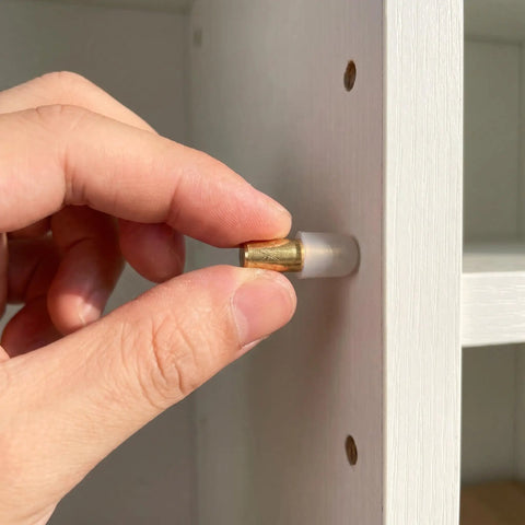 Close-up of hand installing Brass Shelf Support Pegs Set Of 12 With Rubber Sleeves for easy install and precise alignment in white shelf.