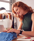 Red-haired woman sewing at a white sewing machine with blue fabric in a garment studio.