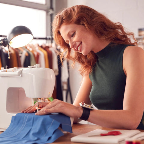 Red-haired woman sewing at a white sewing machine with blue fabric in a garment studio.