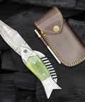 Fish Style Copper Damascus Folding Knife With Ladder Pattern Blade, Brass Engraved Handle, Leather Sheath displayed on black wood