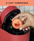Person inserting Fog Chaser Medium Dark Roast K Cup Compatible Coffee Pods 80 Count Single Serve Compostable Pods into red coffee maker