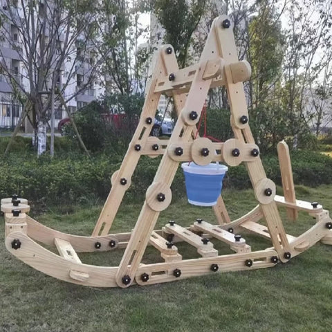 Wooden playground structure with interconnected beams and circular connectors, set up on grass with trees in the background.
