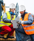 Two workers in vests and helmets discuss plans on a construction site wearing 20 Pack Ratchet Adjustable Safety Hard Hats With Cotton Brow Pad ABS Construction 4 Point Suspension White Bulk Helmets For Men And Women On Job Sites Today