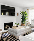 Modern living room with a wall-mounted TV above a fireplace, surrounded by grey and white furniture, houseplants, and natural light.