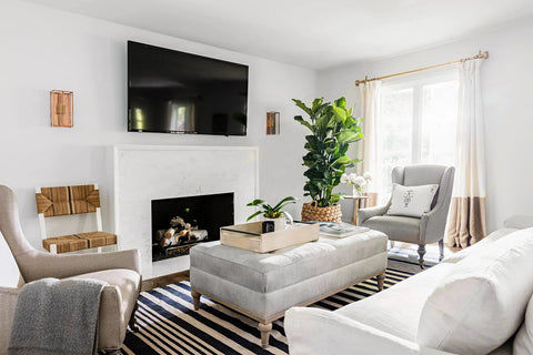 Modern living room with a wall-mounted TV above a fireplace, surrounded by grey and white furniture, houseplants, and natural light.
