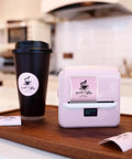 Kickass Portable Bluetooth Label Printer With Slick Wireless Tape Dispenser For Home Office Organization And Shipping Labels No Ink Required Mighty Handy Cordless Thermal Label Maker Handles Wide Continuous Rolls Daily on wooden tray with coffee cup.
