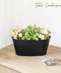 Black flower pot with artificial greenery and white flowers on wooden table, labeled 'Table Centerpiece'.