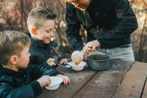 Camping Cookware 18 Piece Mess Kit With Backpacking Stove in use, highlighting anodized pot and serving of food to children outdoors.