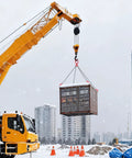 Four-leg steel chain sling with red hooks lifting a shipping container at a snowy construction site from a yellow crane