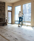 Man standing on ladder applying X Board Paint and Remodel Surface Protector Heavy Duty Breathable Floor Shield in large room.