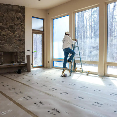 Man standing on ladder applying X Board Paint and Remodel Surface Protector Heavy Duty Breathable Floor Shield in large room.