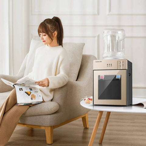 Hot And Cold Tabletop Water Cooler Dispenser For 5 Gallon Bottles, One Button Operation, Food Grade Liner, Perfect For Home, Dorm, Office, Champagne Gold 110V 60Hz next to a seated woman reading a magazine.