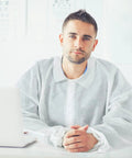 Man wearing 50 Pack Disposable Lab Coats For Adults White Large Bulk With Knitted Cuffs And Collar Ideal For Science Labs Medical Testing Food Prep And Party Themed Events seated at a desk