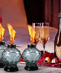 Four Pack Glass Table Torches With Citronella For Outdoor Patio And Garden, Crystal Gem Design, lit beside champagne glasses.