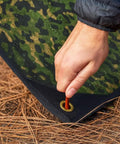 Hand securing Rugged Survival Tarp With Insulated Reflective Mylar Layer for all weather camping with grommet on woodland camo tarp.