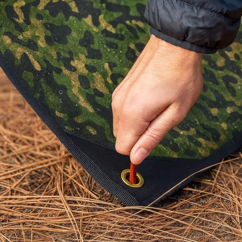 Hand securing Rugged Survival Tarp With Insulated Reflective Mylar Layer for all weather camping with grommet on woodland camo tarp.