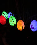 Colorful lit eggs from 20 LED Solar Easter Egg Lights Outdoor Stake With 25 Ft Cord in night garden, featuring bunny and flower patterns.