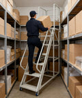 Employee using Badass Heavy Duty Seven Step Rolling Ladder With Reinforced Steel Handrails And Easy Glide Wheels For Warehouse Garage Storage Access 1500 LB Capacity Non Slip Platform to reach high shelves.