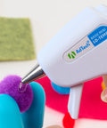 Mini glue gun applying clear glue to purple pompom for crafting.