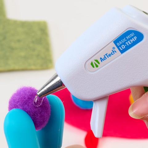 Mini glue gun applying clear glue to purple pompom for crafting.