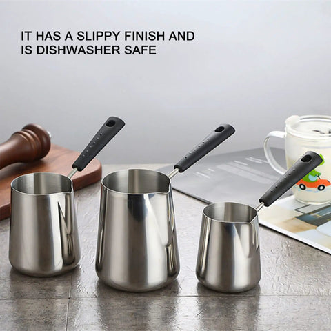 Front view of three milk frothing pitchers in stainless steel with long handles on a kitchen countertop.