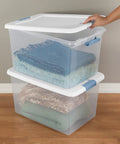 Person stacking 64 Quart Clear Latching Storage Boxes 4 Pack with White Lids, filled with folded blankets on wooden floor.