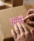 Applying hot pink fragile sticker on box, "Please Handle With Care," part of 500 pack, ultra strong adhesive, easy peel for shipping.