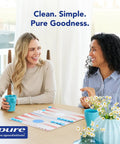 Two women sitting at a table with board game, blue mugs, and branded Pure Encapsulations logo, floral vase in foreground.