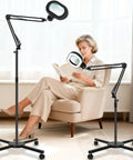 12x Large Floor Magnifying Glass With Light And Stand used by woman reading in a chair, showing adjustable arms and rolling base.