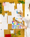 Woman organizing notes and colorful charts on a cork board using Hella Versatile 200 Pcs Push Pins And Heavy Duty Thumb Tacks For Bulletin Board Cork Wall Organizers Featuring Nifty Multi Color Assortments That Are A Solid Go To Choice For Home Office Projects.