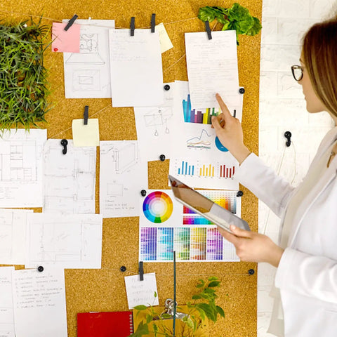 Woman organizing notes and colorful charts on a cork board using Hella Versatile 200 Pcs Push Pins And Heavy Duty Thumb Tacks For Bulletin Board Cork Wall Organizers Featuring Nifty Multi Color Assortments That Are A Solid Go To Choice For Home Office Projects.